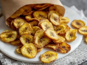 Air Fryer Banana Chips 40 Crispy golden Air Fryer Banana Chips spilling out of a brown paper bag onto a white plate, ready for a healthy snack.