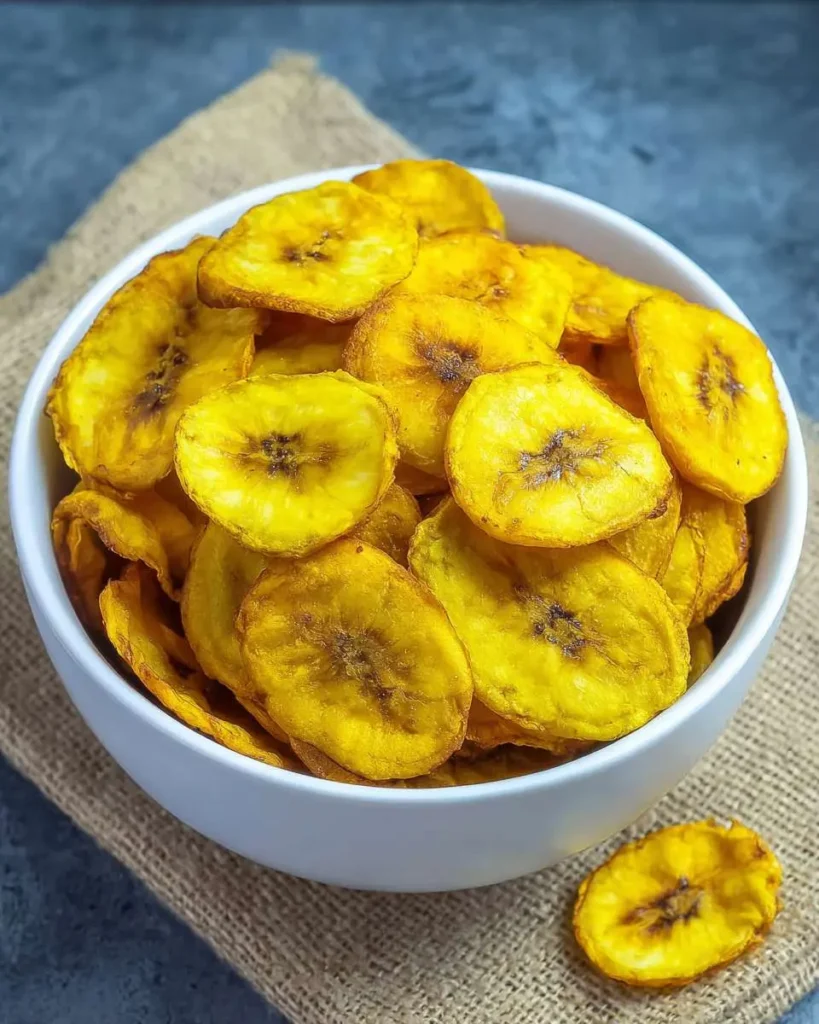 A white bowl filled with golden and crispy Air Fryer Banana Chips, with one chip fallen onto the brown burlap surface.