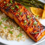 A delicious Air Fryer Gochujang Salmon fillet, glazed and topped with sesame seeds and green onions, served with brown rice, asparagus, and orange slices on a white plate.