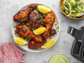 A white plate of deliciously charred air fryer tandoori chicken drumsticks garnished with fresh parsley and lemon wedges, served alongside a green salad and dipping sauce.