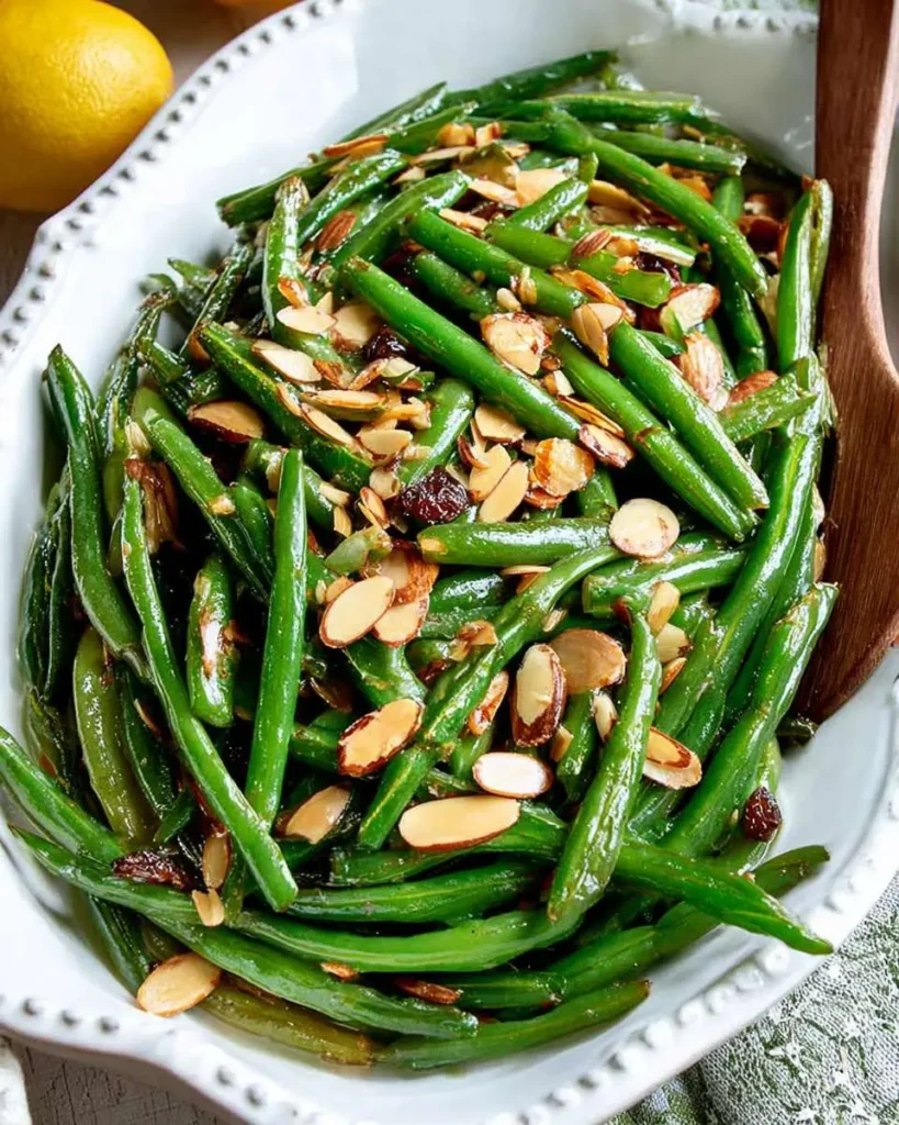 Green Beans Almondine 26 A white serving dish filled with vibrant green beans topped with toasted sliced almonds and brown bits, with a fresh lemon in the top left corner.