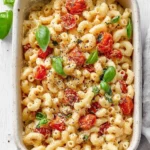A close-up shot of a white casserole dish filled with creamy Baked Feta Pasta, featuring elbow macaroni, roasted cherry tomatoes, and fresh basil leaves.