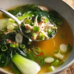 A spoon scoops shredded chicken and green onions from a steaming bowl of delicious bok choy chicken soup with tender greens.