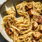 A close-up of a fork in a bowl of creamy cowboy butter chicken linguine, featuring grilled chicken pieces and herbs.