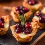 Close-up of golden puff pastry Cranberry brie bites with melted cheese, cranberry topping, and rosemary garnish on a slate board.