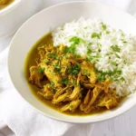 A white bowl holds flavorful curry chicken alongside fluffy white rice, garnished with fresh herbs.
