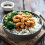 A delicious bowl of honey garlic shrimp with white rice and steamed broccoli, garnished with sesame seeds on a wooden table.