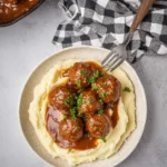 A comforting bowl of mashed potatoes topped with savory meatballs and gravy, garnished with fresh parsley, next to a pan.
