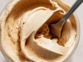 A spoon scoops creamy Ninja Creami Cinnamon Ice Cream from a clear tub, revealing its smooth texture and speckled cinnamon dust.