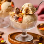 A spoon scoops a bite from a glass dish of creamy Ninja Creami Peanut Butter Cup ice cream, topped with mini peanut butter cups and candies.