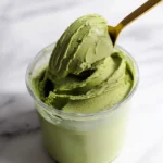 A golden spoon lifts a scoop of vibrant green Protein Matcha Ninja Creami from a clear container, resting on a white marble surface.