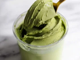A golden spoon lifts a scoop of vibrant green Protein Matcha Ninja Creami from a clear container, resting on a white marble surface.
