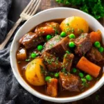 A close-up of a hearty slow cooker beef stew in a white bowl, filled with tender beef, potatoes, carrots, and green peas, garnished with parsley.
