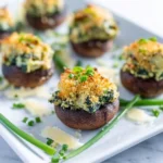 Close-up of a white platter featuring several golden-brown spinach artichoke stuffed mushrooms, garnished with fresh chives and parmesan shavings.