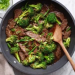 A skillet of savory steak and broccoli stir fry garnished with green onions and red pepper flakes, with a wooden spoon.