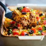 A serving of a cheesy Taco Casserole with ground beef, tomatoes, olives, and green onions being lifted from a baking dish.