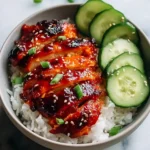 A bowl of white rice topped with glistening sliced Air Fryer Gochujang Chicken Thighs, sprinkled with sesame seeds and green onions, accompanied by fresh cucumber slices.