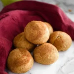 A pile of golden brown almond flour rolls, lightly dusted with cinnamon, rests on a white marble counter next to a red cloth.