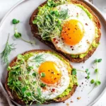 Two pieces of avocado toast with egg, topped with vibrant microgreens and red pepper flakes, served on a white plate.