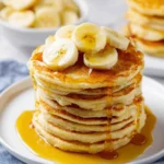 A stack of golden banana pancakes generously drizzled with maple syrup and topped with fresh banana slices, with a bowl of bananas in the background.