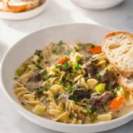 A steaming bowl of creamy beef noodle soup with egg noodles, beef chunks, carrots, celery, and parsley, served with crusty bread.