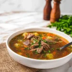 A close-up of a hearty beef short rib soup in a white bowl, filled with tender meat, potatoes, carrots, and fresh herbs, served on a textured placemat.