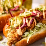 A close-up of a delicious Bird Dog Sandwich featuring crispy fried chicken, shredded lettuce, red onion, bacon, pickles, and a creamy sauce in a hot dog bun.