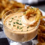 A golden fried onion ring is dipped into a clear glass bowl of creamy Blooming Onion Sauce, garnished with parsley.