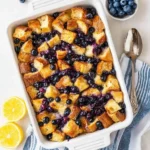 A top-down view of a golden brown Blueberry French Toast Casserole in a white baking dish, garnished with fresh blueberries and lemon halves.