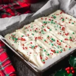 A baking pan filled with frosted Christmas Sugar Cookie Bars, topped with red, green, and white sprinkles, next to a plaid cloth and holly.