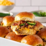Close-up of hearty Cowboy Sliders stacked in a white dish, revealing ground beef, melted cheese, crispy bacon, and sliced jalapeños.