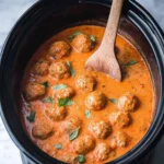 A black crockpot filled with creamy, orange Crockpot Garlic Parmesan Meatballs garnished with fresh basil and a wooden spoon.