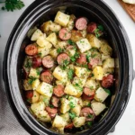 Overhead view of a black crockpot filled with cooked Crockpot Potatoes and Kielbasa, seasoned and garnished with fresh parsley.