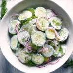 Refreshing Cucumber Yogurt Salad in a white bowl, with sliced cucumbers, red onions, creamy dressing, fresh dill, and black pepper.