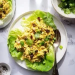 A vibrant plate of Curry Chicken Salad served in crisp lettuce cups, garnished with cilantro and raisins, on a marble table.