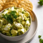 A bowl of creamy Egg Avocado Salad topped with fresh parsley and black pepper, served with crispy crackers on a white plate.