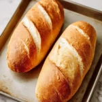 Two freshly baked golden-brown loaves of French Bread with characteristic diagonal slashes resting on a rustic metal baking sheet.