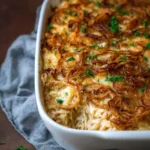 A close-up of a French Onion Chicken Orzo Casserole in a white baking dish, topped with golden crispy fried onions and fresh parsley.