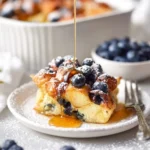 A close-up of a slice of Hawaiian Roll French Toast Bake, generously topped with fresh blueberries, powdered sugar, and maple syrup pouring over it.