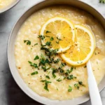 A close-up shot of a creamy bowl of lemon rice soup garnished with fresh lemon slices, chopped parsley, and thyme sprigs.