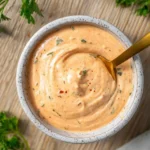 A top-down view of creamy, orange Louisiana Remoulade Sauce in a speckled bowl with a golden spoon, garnished with green herbs.