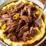 A hearty bowl of tender Mississippi Pot Roast shredded over creamy mashed potatoes, topped with gravy and pickled peppers, with a fork.