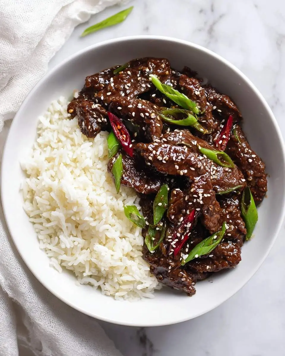 Mongolian Beef 27 A white bowl filled with glistening Mongolian Beef, fluffy white rice, topped with sesame seeds, green onions, and red chili slices.