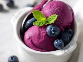 Two scoops of vibrant purple Ninja Creami Blueberry Frozen Yogurt topped with fresh blueberries and a mint leaf in a white bowl with a spoon.