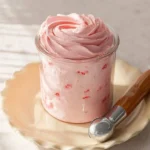 A glass jar filled with a vibrant pink Ninja Creami Strawberry Frozen Yogurt, topped with creamy swirls, on a ruffled plate with an ice cream scoop.