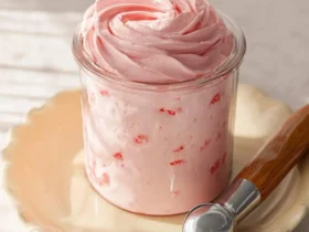 A glass jar filled with a vibrant pink Ninja Creami Strawberry Frozen Yogurt, topped with creamy swirls, on a ruffled plate with an ice cream scoop.