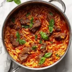 One Pot Beef Giouvetsi in a stainless steel skillet, featuring tender beef, orzo pasta, and fresh basil in a rich tomato sauce.