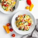 Two bowls of vibrant Greek-inspired orzo salad with tomatoes, cucumbers, feta, and olives on a white table with fresh ingredients.