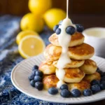 A stack of golden brown pancake poppers topped with fresh blueberries and a drizzle of white sauce, with lemons in the background.