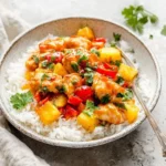 A close-up of a bowl filled with white rice, topped with sweet and sour pineapple chicken, red bell peppers, and fresh cilantro, with a fork.